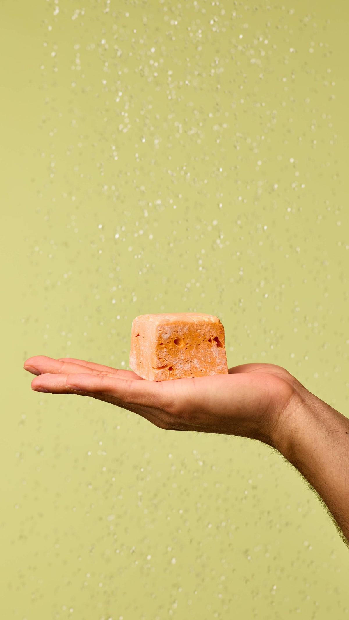 The image shows a warm-toned hand holding a pastel orange Passion Fruit Delight floating soap, resembling a dense marshmallow. Water droplets fall around the hand and soap, against a light green background.