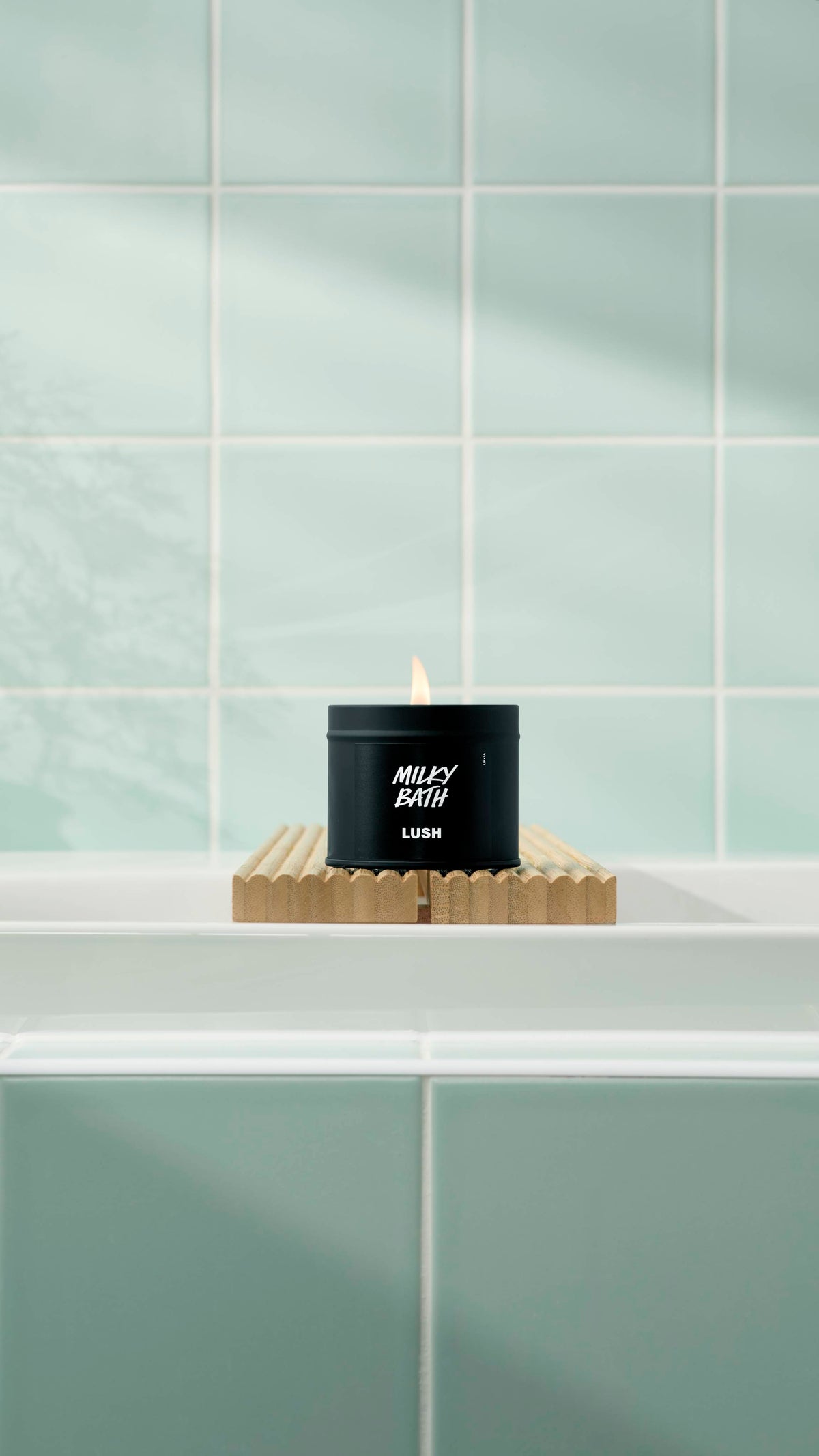 A lit Milky Bath candle in a matte black tin sits on a fluted wooden bath bridge over a white tub. The background features soft mint green square tiles with a gentle shadow of a plant.