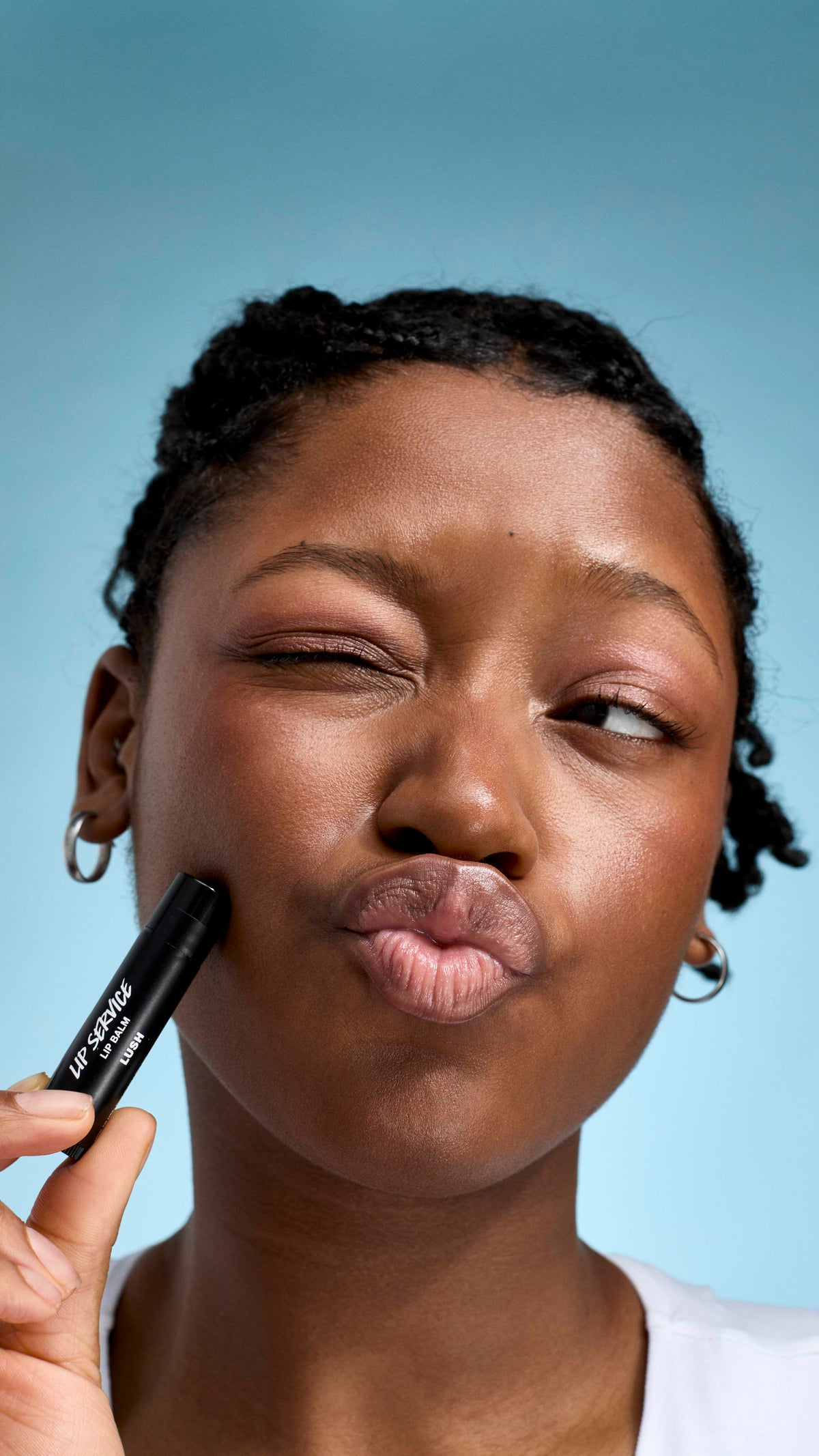 A close-up of a model with braided hair and glowing skin, making a playful pouting face with one eye shut. They hold a black tube of Lush 'Lip Service' lip balm against their cheek, set against a soft, light-blue background.