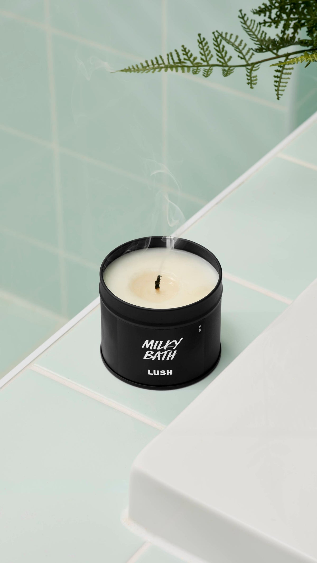A close-up, high-angle shot of an extinguished Milky Bath candle by Lush in a matte black tin. Thin wisps of smoke rise from the blackened wick. It sits on a white ledge against mint green wall tiles with a fern leaf visible at the top.