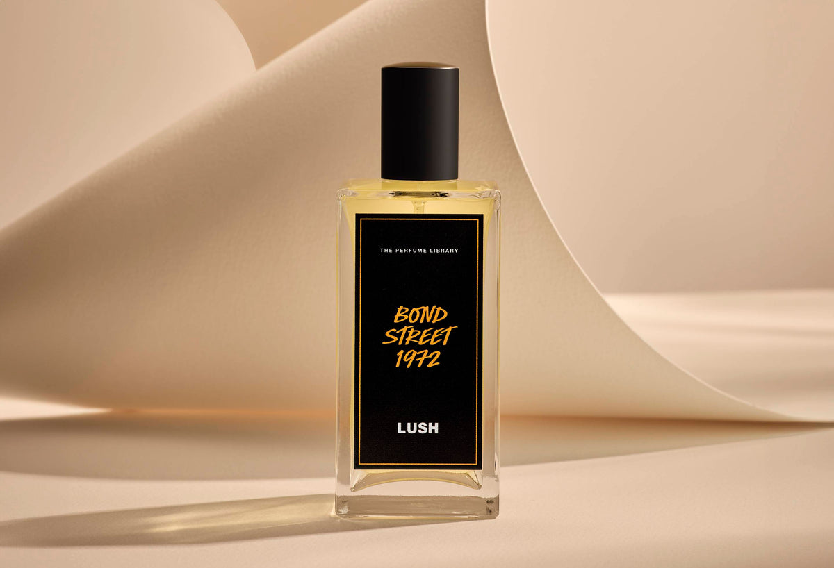 The image shows the Bond Street 1972 glass perfume bottle. It is sitting on a soft, beige background with curved, cream paper creating soft, gliding shapes behind the product.